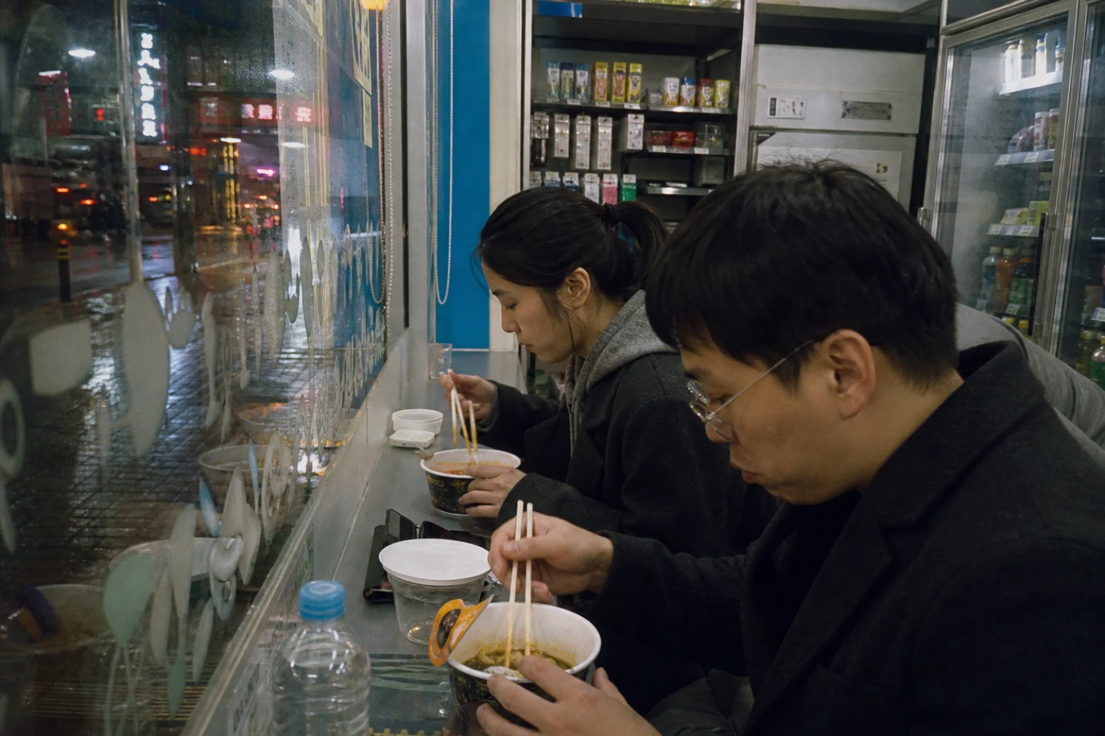 Two people eating a simple late-night meal at a neighborhood convenience store window counter