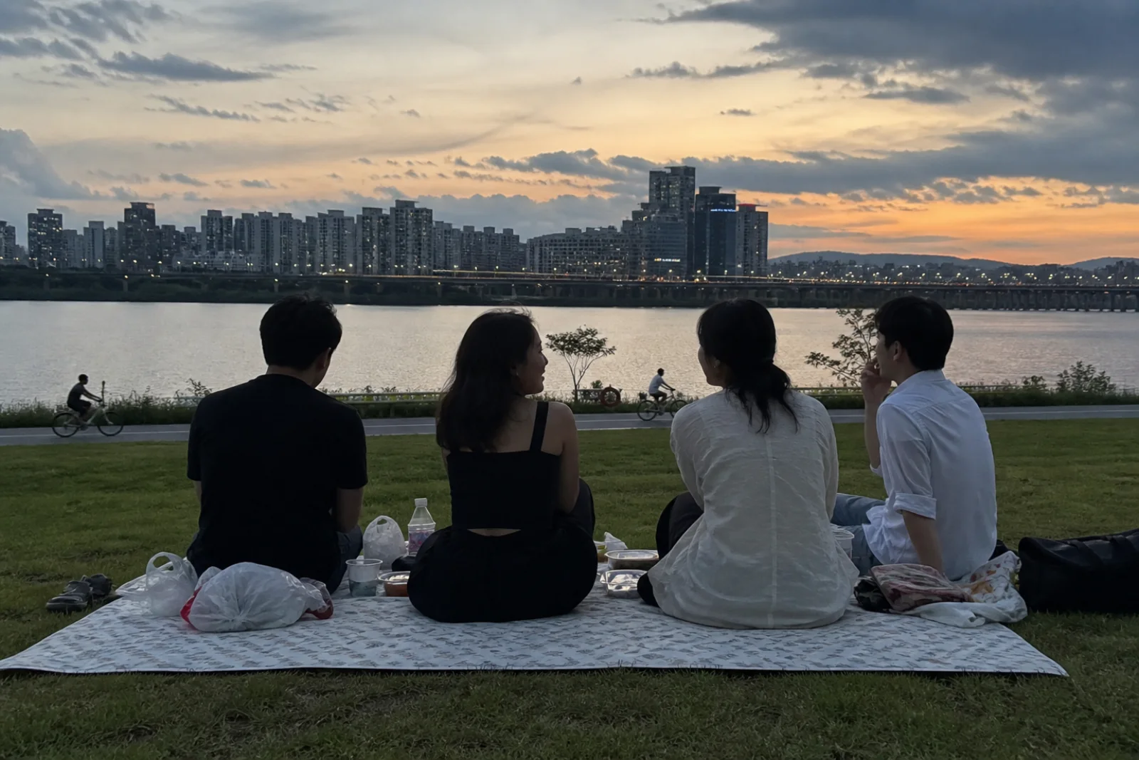 Friends sitting on a simple picnic mat beside the Han River at dusk