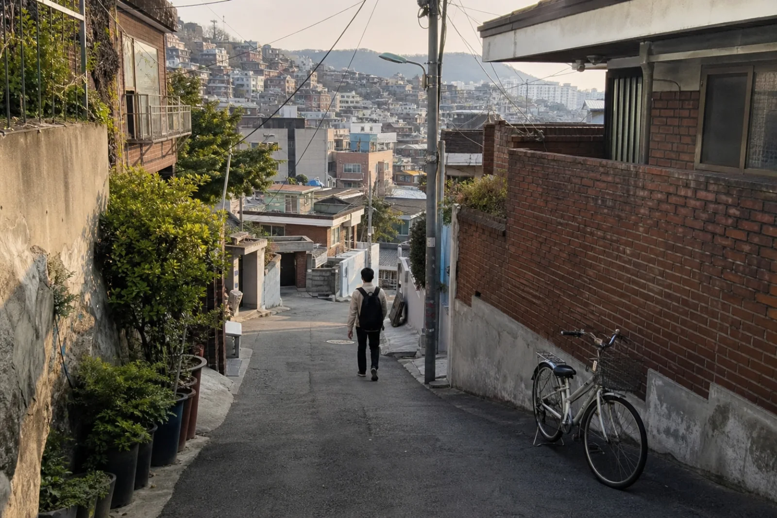 A person strolling through a quiet, lived-in Seoul hillside alley in late afternoon