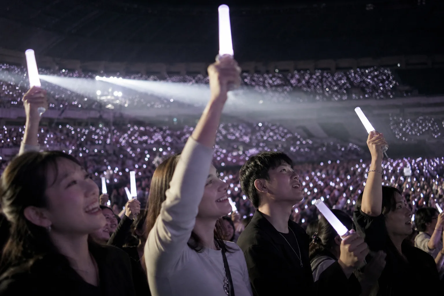 Fictional adult K-pop fans raising plain glowing light sticks together inside a crowded concert arena
