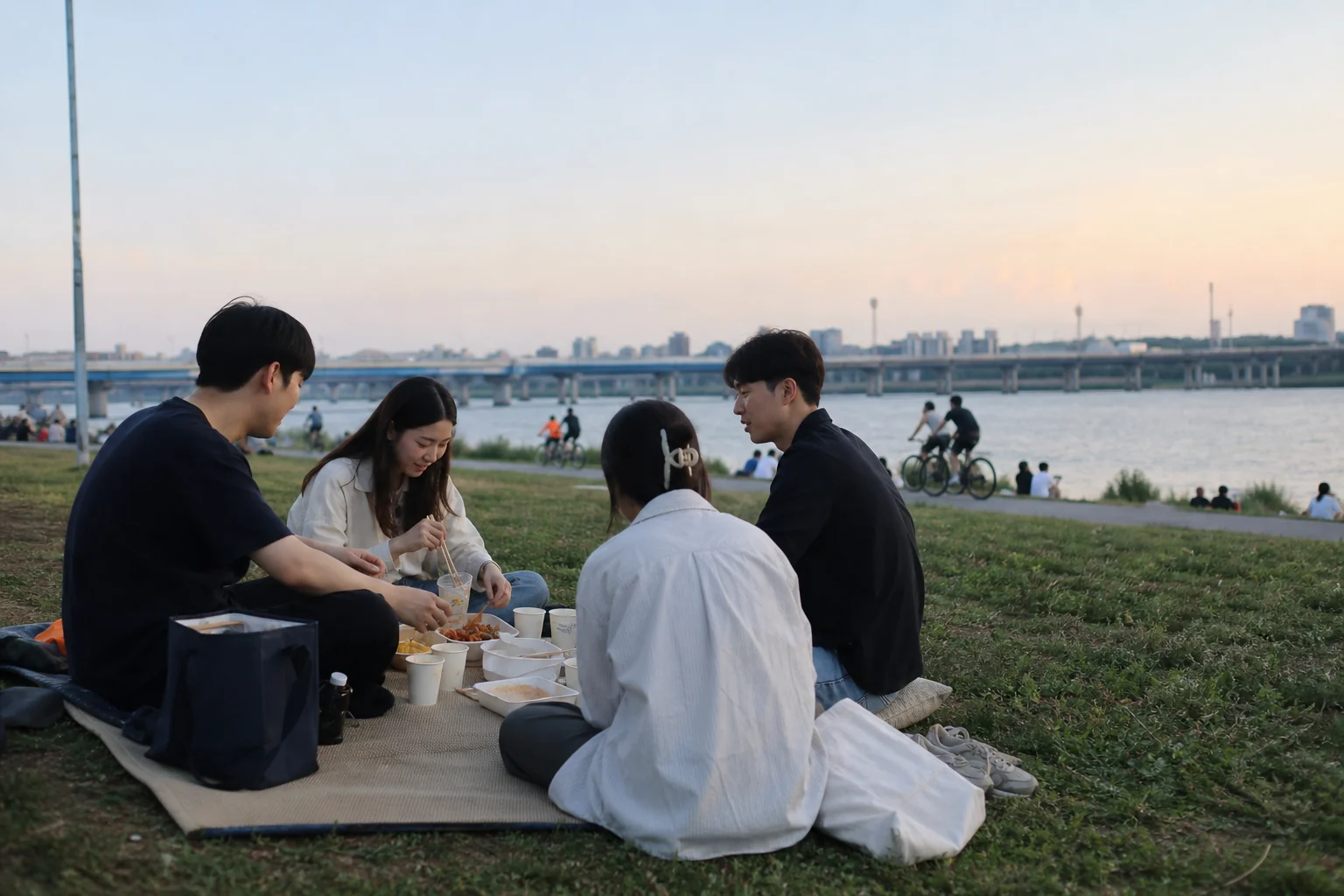 Four fictional friends sharing an evening picnic on grass beside the Hangang while cyclists pass behind them