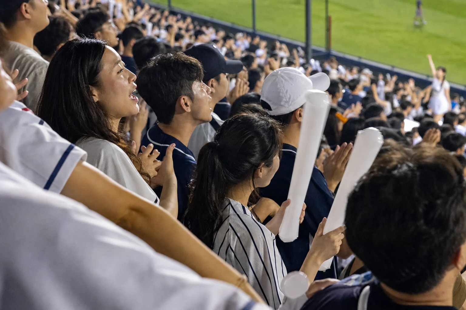Fictional Korean baseball fans singing and clapping with plain cheering sticks during an evening game