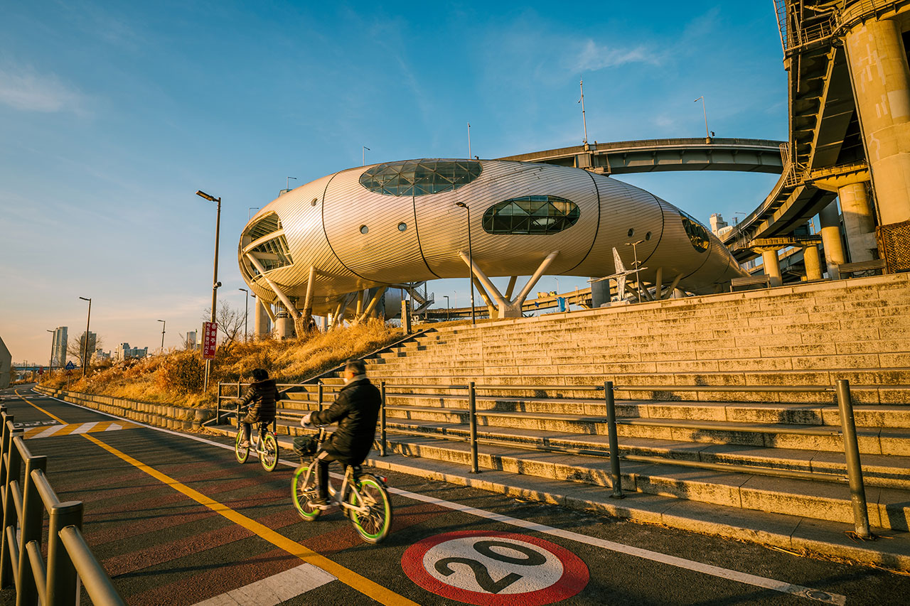 Two cyclists passing the silver J-Bug cultural structure beside the bicycle path at Ttukseom Hangang Park