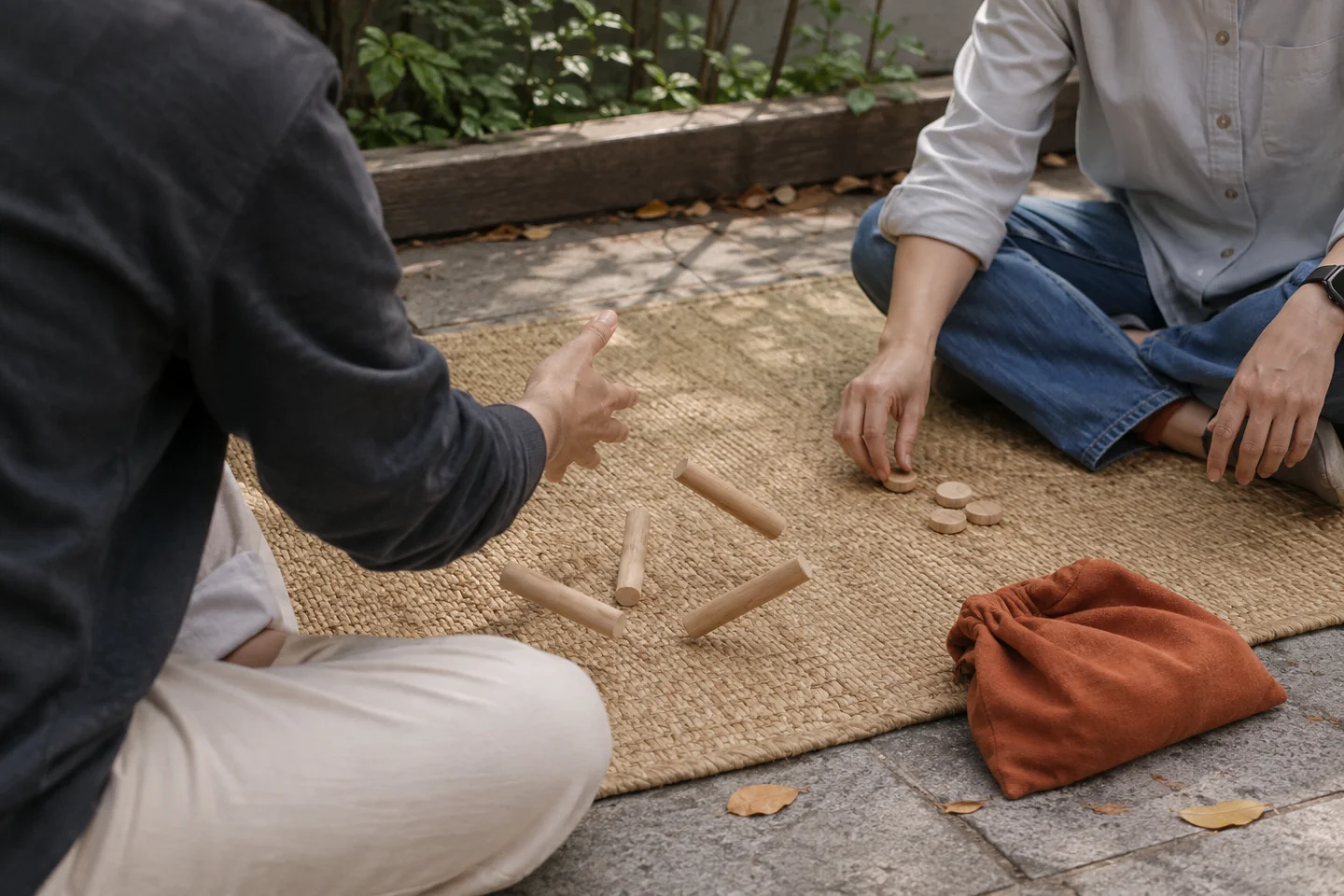 Two anonymous adults sit on a woven mat and play with four wooden yut sticks in a fictional courtyard