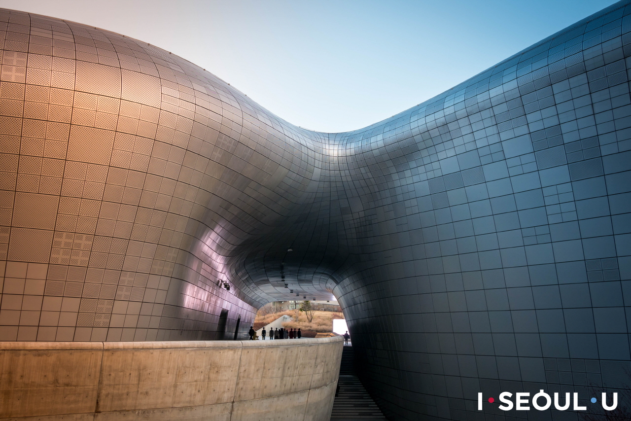 Sunlit curved metal passage at Dongdaemun Design Plaza with a small group of visitors in the distance