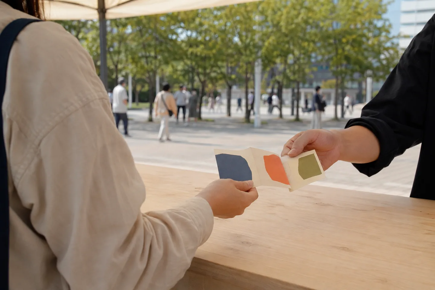 Two anonymous adults pass a blank accordion-fold paper object across a plain outdoor market table