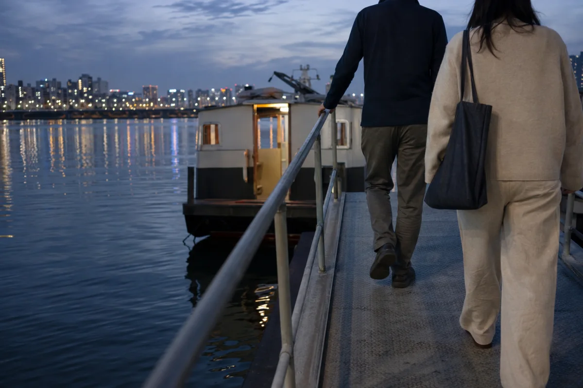 Two anonymous adults walk down a plain river-pier gangway toward a small unbranded ferry at blue hour