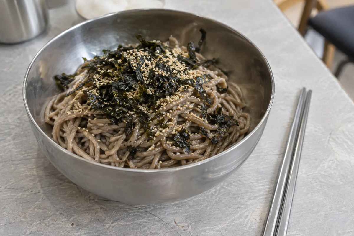 Oil-dressed grey-brown buckwheat noodles with seaweed and sesame in a steel bowl on a light tabletop.