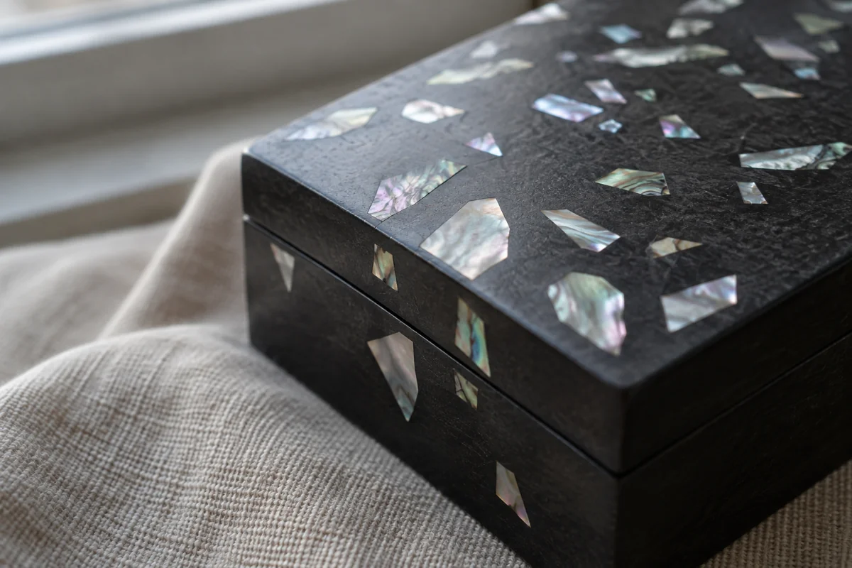Imagined dark lacquer-style box with small iridescent shell inlays in an abstract arrangement, photographed close up; AI illustration, not an actual museum object or product.