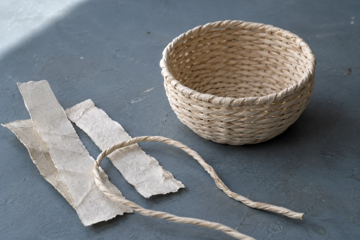 AI-generated editorial illustration of an invented ivory paper-cord bowl beside blank paper strips and a loose twisted cord on a blue-gray table; not a documented craft object.